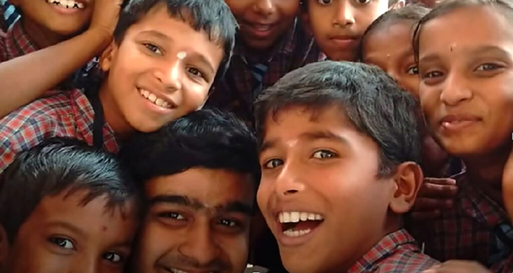 Smiling schoolchildren in checked uniforms crowd close together, with a boy in the foreground laughing at the camera.
