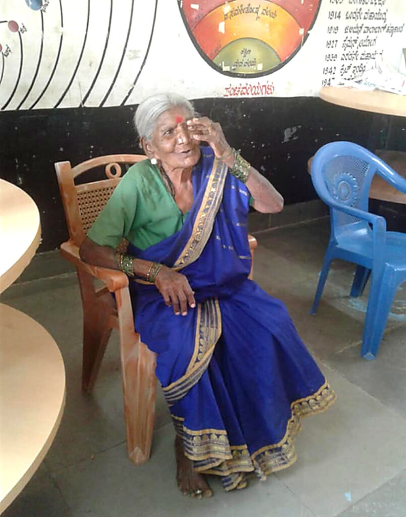 An elderly woman in a blue saree and green blouse sits in a wooden chair, with one hand raised to her forehead.