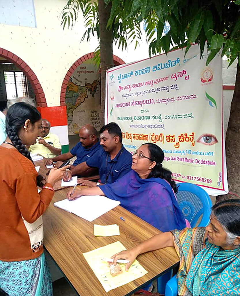 Volunteers and staff sit at a table registering villagers at an eye-care camp, with a banner behind them.