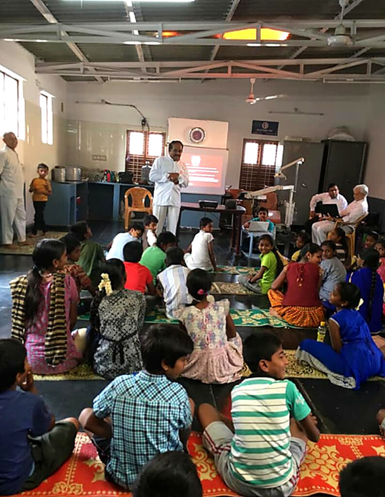 Children sit on colourful mats in a hall, listening to a man in white who is speaking in front of a screen.