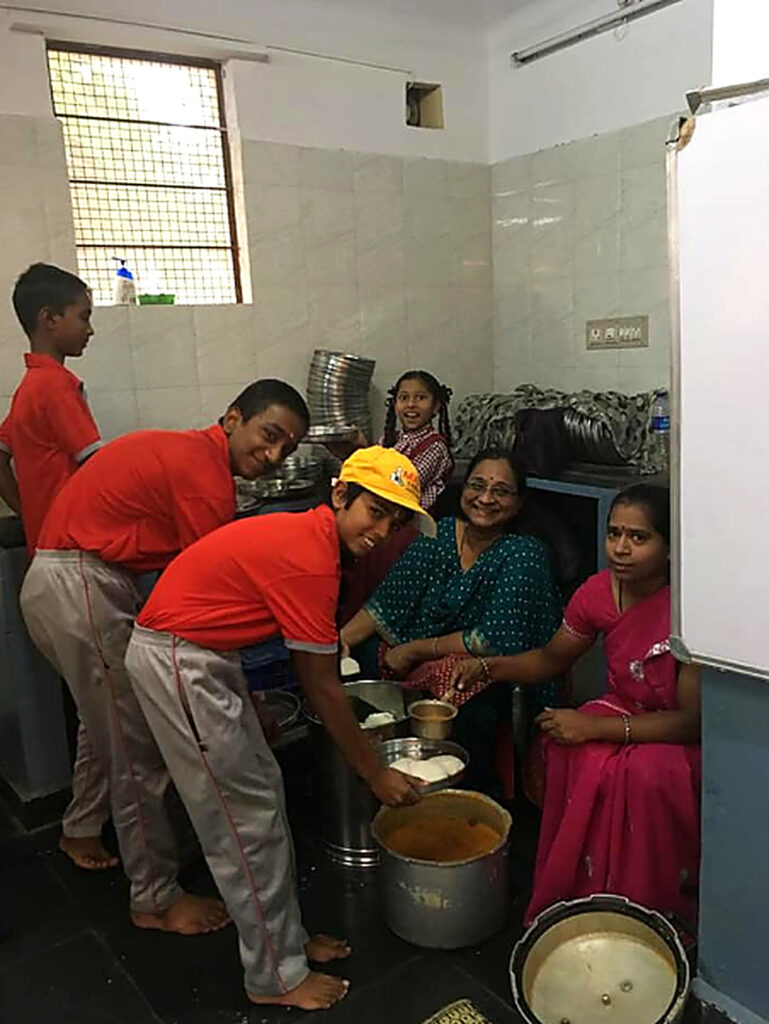 Boys in red T-shirts, a girl and two women prepare food together in a kitchen with large pots and steel plates.
