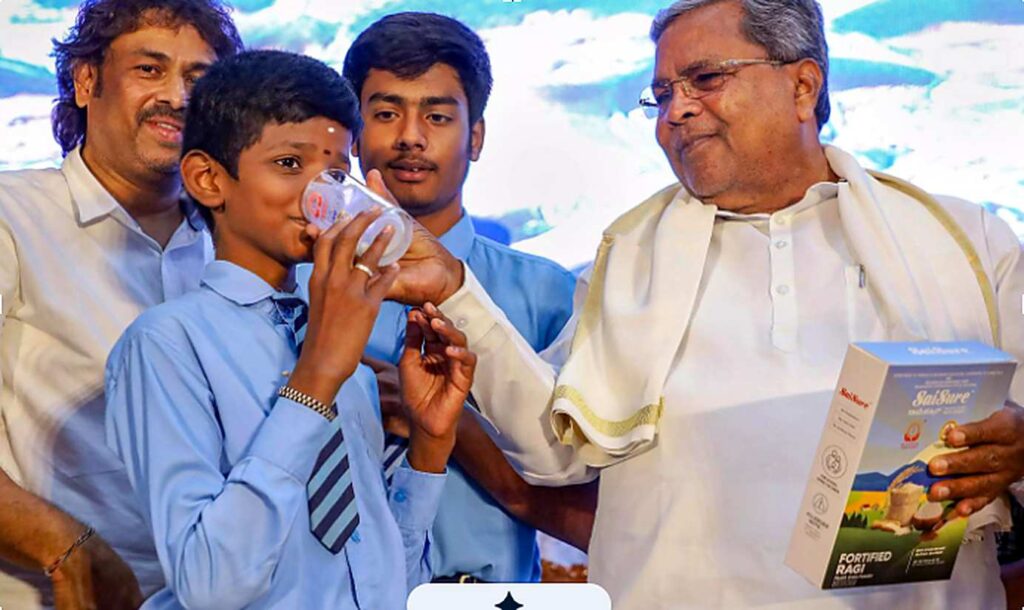 A schoolboy in a blue uniform drinks from a glass at a stage event while an elderly man in a white shirt and shawl holds a box of SaiSure Fortified Ragi and smiles at him. Two other people stand behind them.